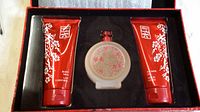Three-piece fragrance set displayed in black velvet-lined red satiny floral print box, includes perfume spray bottle center, Body Milk tube left, Shower Gel tube right.
