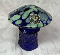 Close-up image of dark blue and green hand blown glass mushroom with Golden Crown E and R label on top.