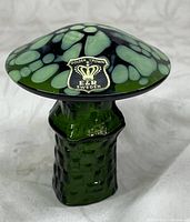 Close-up of the hand blown glass mushroom showing the cap with green and black colors intermixed with light green spots and the original Golden Crown E & R Sweden label affixed.