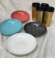 Four colorful Branchel melamine bowls in white, blue, coral pink, and dark gray with four gold-tone Therma-Ware insulated tumblers with black tops arranged on a white fabric.