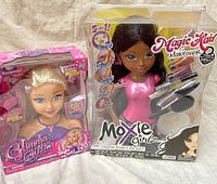 Two boxed fashion doll styling heads: a Moxie Girlz Magic Hair Makeover and a Glimma Girlz Style My Hair, showing front of packaging with doll faces and included hair styling accessories.