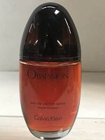 Front view of Calvin Klein Obsession eau de parfum spray bottle with cap on, showing brown amber liquid and label text.