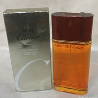 Photo of Must de Cartier 3.3 oz Eau de Toilette bottle with amber liquid and dark red cap, alongside reflective silver box with 'Must de Cartier' text on front.
