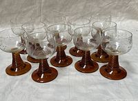 Full view of 12 etched crystal barware glasses with amber stems and bases arranged on white surface.
