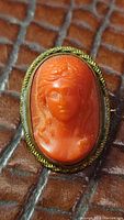 Close-up image showing detailed carving of the coral cameo depicting a woman's profile with intricate facial features and hair, set in a metal frame with gold twist wire detailing.