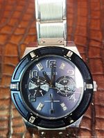 Front view of Guess men's wristwatch showing black dial with crystal markers, subdials, and silver metal band on textured brown surface.