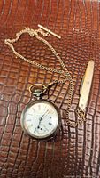 Photo showing the pocket watch with attached gold-filled chain and accessory on a brown textured surface.