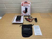 Photo of packaged and unpacked components laid out on table: jump starter, jumper cables, USB cable, pouch, manual, and product box.