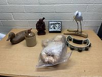 Photo showing the assortment of items on a table against white brick background including wooden duck, ceramic jug, Buddha statue, photo frame, bird sculpture, tambourine, and baby doll in plastic bag.