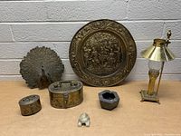 Wide view of all items in lot including brass peacock figurine, decorative brass plate, two small brass perforated boxes, brass table lamp, small stone animal figurine, and dark ashtray
