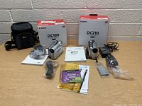 Photo showing both Canon camcorders with their boxes, cables, manuals, batteries, disc media and black camera bag on table.