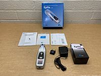 Photo showing Epila SI-808 hair removal device, manuals, power adapter with plug, and two boxes arranged on a wooden surface against a white brick wall.