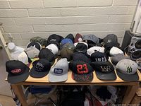 Wide view of 24 baseball style caps arranged on a wooden table against a beige brick wall. Various colors including black, white, grey, navy, and camouflage. Some caps have logos or text.