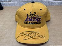 Yellow baseball cap bearing Los Angeles Lakers logo with embroidered crown and word 'Champion', signed on brim with black marker.