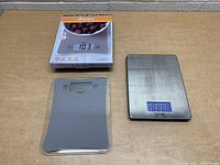 Two digital food scales shown on a table. One scale is in original Allrecipes box showing a transparent platform and digital display. The other is a stainless steel Taylor scale with digital readout lit blue.