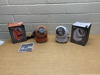 Two desk fans with original packaging and manual placed on a table with a brick wall background.