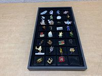Overview of 27 assorted enamel pins in black display tray showing various characters and brand logos