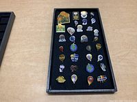 Image showing 27 assorted enamel ballooning pins laid out on a black felt tray arranged in rows.