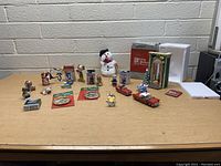 Wide view of the Christmas ornament lot featuring Looney Tunes characters, vintage cars, a snowman figure, small trees, and packages of pins and ornaments on a table against a white brick background.
