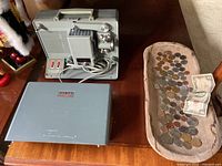 Overview of Argus M-500 film projector and its cover on a wooden surface next to a wooden bowl filled with various foreign coins and a US $2 bill.