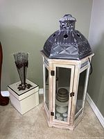 Full view of lantern-style candle holder with metal top, glass panels, several tealights inside, a decorative item inside, a nearby tall etched glass vase, and a white storage box.