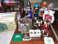 Photo showing the full lot of wooden nutcrackers, Dickens Globe Theatre Village building, original box, booklet, and figurines in packaging arranged on floor near chair