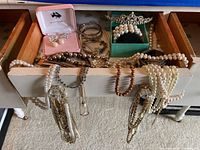 Wide view of open drawer showing assortment of faux pearl and stone necklaces, silver-tone bangles, earrings in pink box, and tiara in green box.