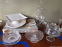 Overview of multiple glass cake plates, platters, candy dishes, and ceramic platters and cake dishes shown on a table with boxes underneath.