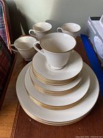 Stacked Lenox Heyworth china including teacups, saucers, dessert plates, salad plates, and dinner plates, all cream-colored with gold trim.