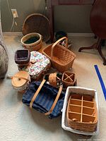 Overview of eleven woven baskets and accessories on the floor including rectangular, round, and fabric-lined pieces, plus a blue plaid insulated bag and wooden butter dish.
