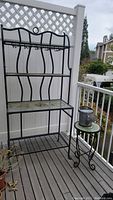 Full view of metal planting stand, watering can on tile top side table on deck with white fence background