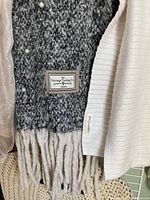 Close up of gray and cream knitted poncho with The Heritage Trading Co. label and Calaberry Creek tag, pearl button embellishments, and yarn tassels