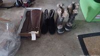 Three pairs of women's boots on floor: tall brown Lamo boots in box, black Franco Sarto boots, and Sorel lace-up snow boots with faux fur trim