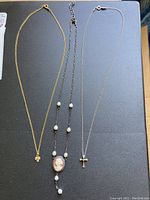 All three necklaces laid flat on a dark surface showing gold filled chain with leaf cluster pendant, sterling silver chain with cross pendant and longer sterling silver cameo necklace with faux pearls.
