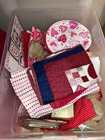 Clear plastic bin with folded Valentine's Day themed textile pieces in red, pink, white, and heart patterns, a round plate with heart motifs, and plush teddy bears inside.