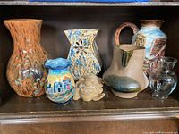 Seven ceramic and glass vases and one ceramic face statuette displayed together, showing variety in shapes, colors, and designs.