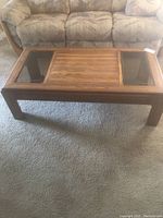 Front view of rectangular wooden coffee table with two glass inset panels on the surface, showed against a carpet and couch background.
