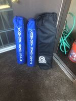 Three folding camp chairs in carry bags placed upright near a door and garden hose. Two blue bags labeled 'GreatLand Comfort Quad Chair', one black bag labeled 'Yukon Charlie'.