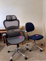 Two office chairs positioned side by side near an office desk: a black mesh rolling armchair with headrest and a blue padded rolling side chair.