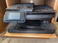 Front view of black HP Photosmart 7525 printer showing touchscreen, paper tray and scanner lid with dust and signs of storage.