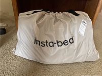 Beige drawstring carry bag with 'insta-bed' printed on front, placed under a wooden desk on carpeted floor.