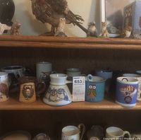 Shelf with 13 ceramic mugs, mostly owl-themed designs. Includes a turquoise owl mug with spoon, brown owl mug, white mug with blue owl drawings, and other assorted mugs.