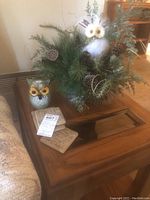 Photo showing owl arrangement with greenery, pinecones, artificial owl and decorative coasters on wooden table.