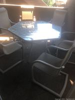 Hexagonal glass-top patio table with six light beige sling armchairs arranged around it, placed on outdoor concrete or patio surface.