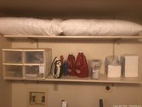 Wide view of utility room shelf with pillows, cleaning bottles, plastic container with drawers, napkins, and light bulbs