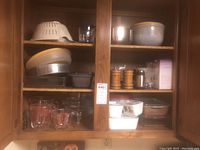View of wooden kitchen cabinet interior shelves showing vintage Pyrex and Corningware glass bowls, measuring cups, plastic colander, and baking dishes.
