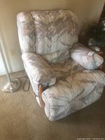 Upholstered recliner chair with patterned fabric and wooden arm accents shown from the left front angle, resting on carpet near a lamp.