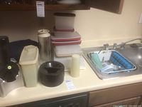Kitchen countertop with stacked Tupperware containers, manual juicer, opaque containers, and sink drainer with dish drying rack beside the kitchen sink.