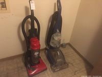 Photo showing the red Dirt Devil upright vacuum cleaner and blue Hoover SpinScrub upright carpet cleaner side by side in a room corner.