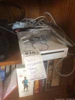 White Nintendo Wii console with cables on top, Wii Sports game case, and DVDs visible in background on shelf.